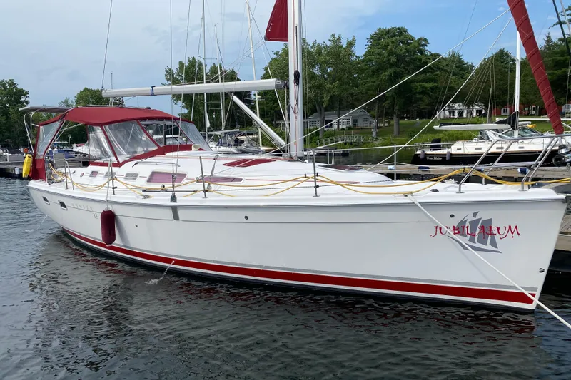 The Image of 2005 Hunter 38 sailboat docked, featuring red accents and canopy, with lush greenery in the background. - 0