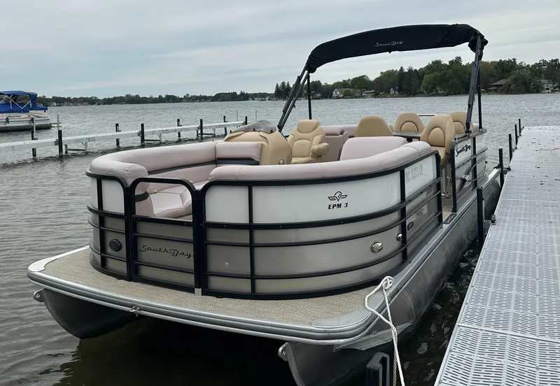 Slide: The Image of A 2019 South Bay pontoon boat docked. It features beige seating and a black canopy. - 1