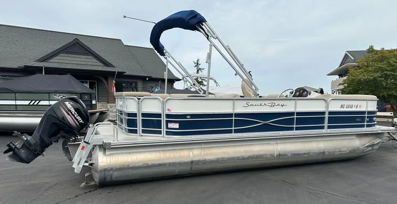 Slide: The Image of A 2018 South Bay S224CR pontoon boat with a blue canopy and Mercury outboard motor, parked on pavement. - 19