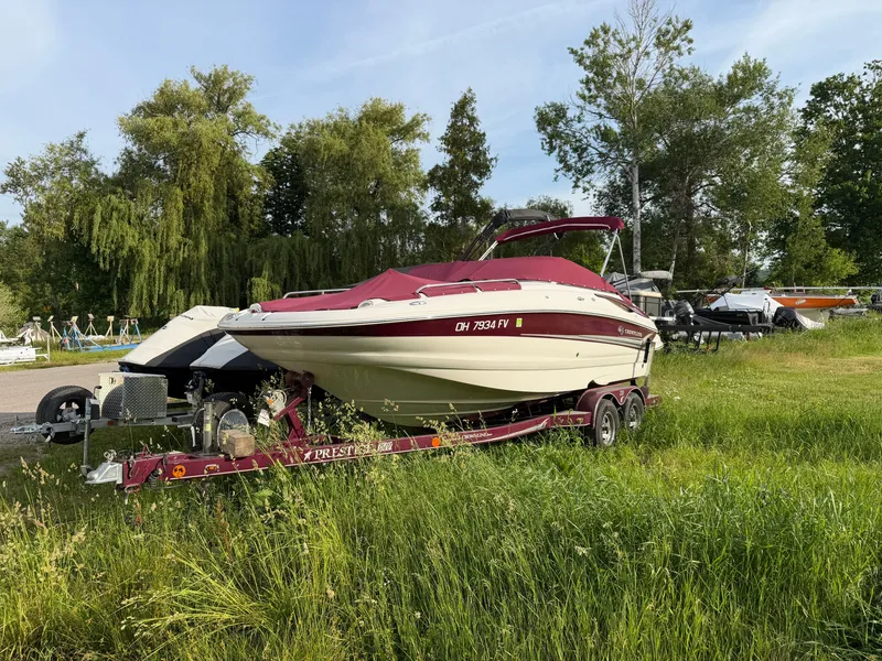 The Image of A 2006 Crownline 220 EX boat on a trailer in grass, surrounded by trees. - 0