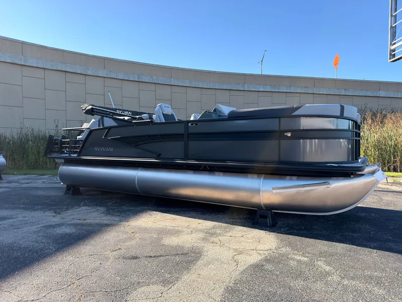 The Image of A modern Sylvan X3 LZ DH pontoon boat is parked on a lot in front of a building. - 0