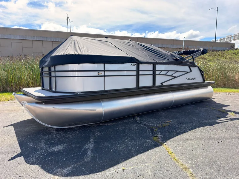 Slide: The Image of A 2027 Sylvan Q-22 ULZ pontoon boat with a black cover, parked on a paved area. - 1