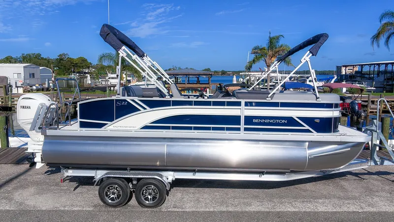 Slide: The Image of A 2027 Bennington 22 S Swingback pontoon boat on a trailer, set against a scenic waterfront. - 22
