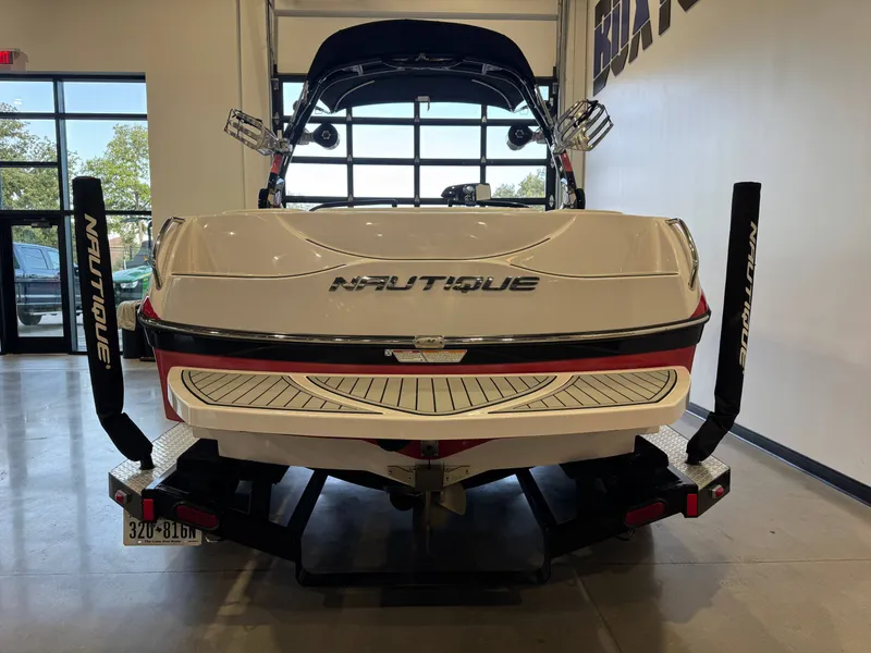 Slide: The Image of The image shows the rear of a white 2011 Nautique Super Air Nautique 210 boat on a trailer indoors. - 6