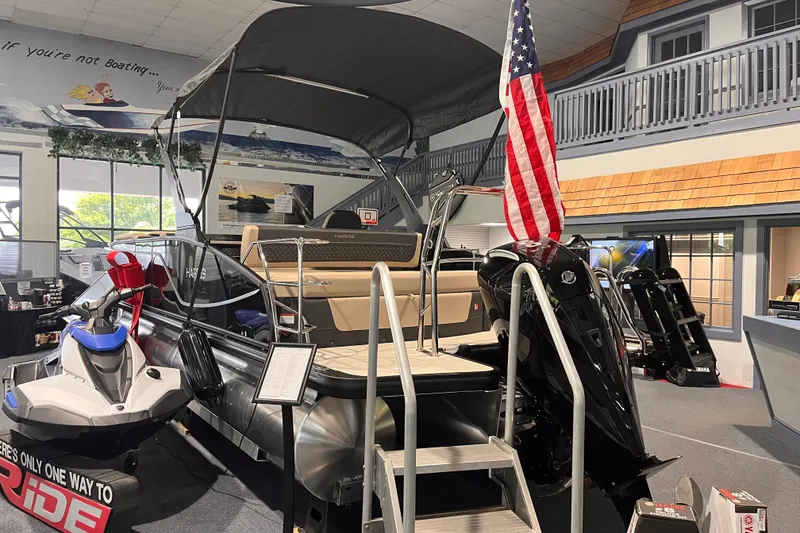 Slide: The Image of 2026 Harris Sunliner 250 pontoon boat displayed indoors with American flag and jet ski. - 2