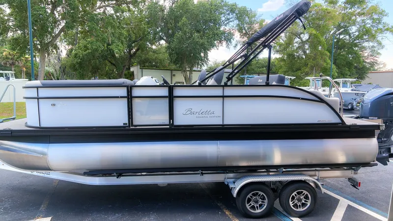 Slide: The Image of The image shows a Barletta SANZA 22UC pontoon boat on a trailer, surrounded by trees. - 5