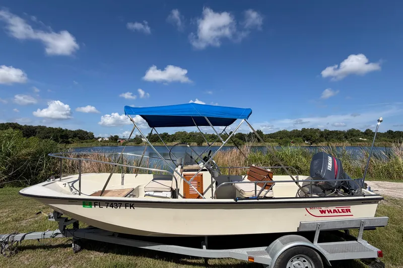 The Image of 1987 Boston Whaler 17 Montauk boat with blue canopy on trailer by a lake. - 0