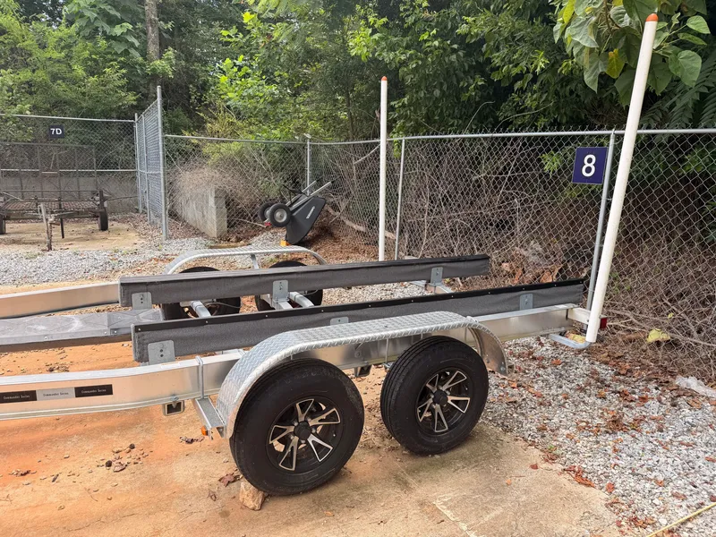 Slide: The Image of A dual-axle boat trailer is parked on a gravel lot, surrounded by trees and a fence. - 44
