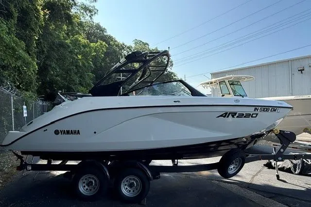 Slide: The Image of 2023 Yamaha Boats AR220 on trailer, parked outdoors near trees and a building. - 2