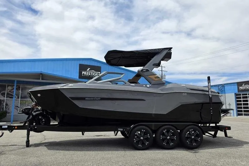 Slide: The Image of 2027 MasterCraft X23 boat on trailer, parked outside a blue building under a cloudy sky. - 0