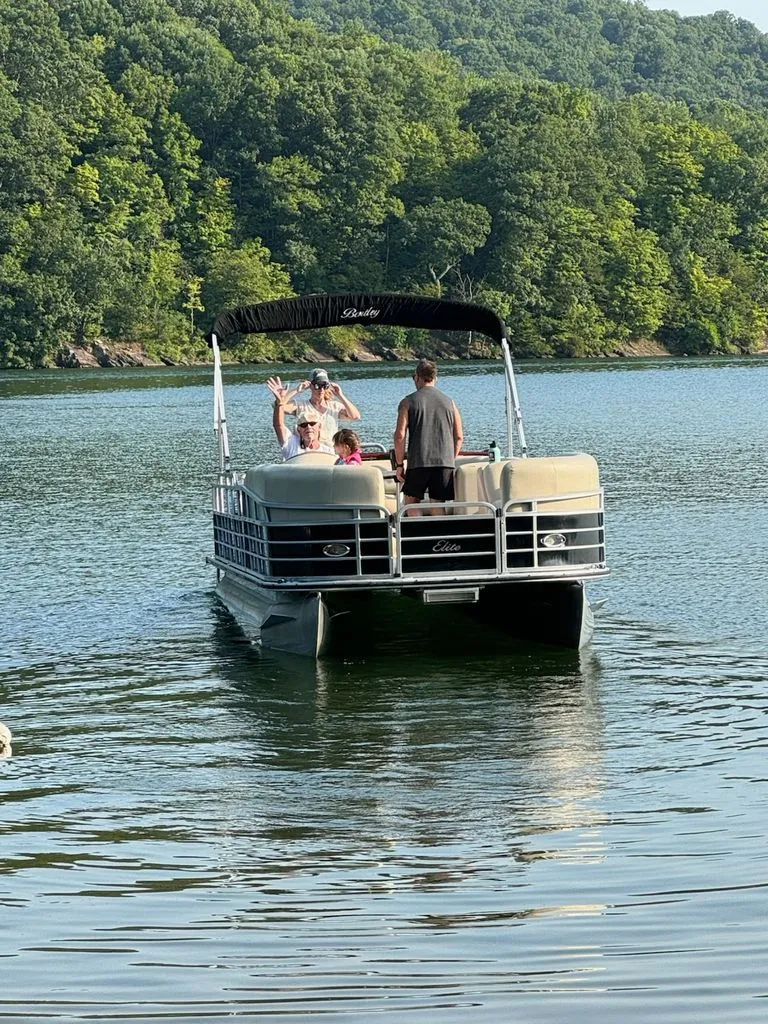Slide: The Image of A 2014 Bentley Pontoons Elite 25 on a calm lake with green trees in the background. - 2