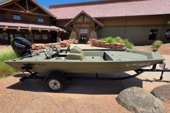 Slide: The Image of 2024 Tracker Grizzly 1754 SC boat on trailer, parked outside rustic building. - 2