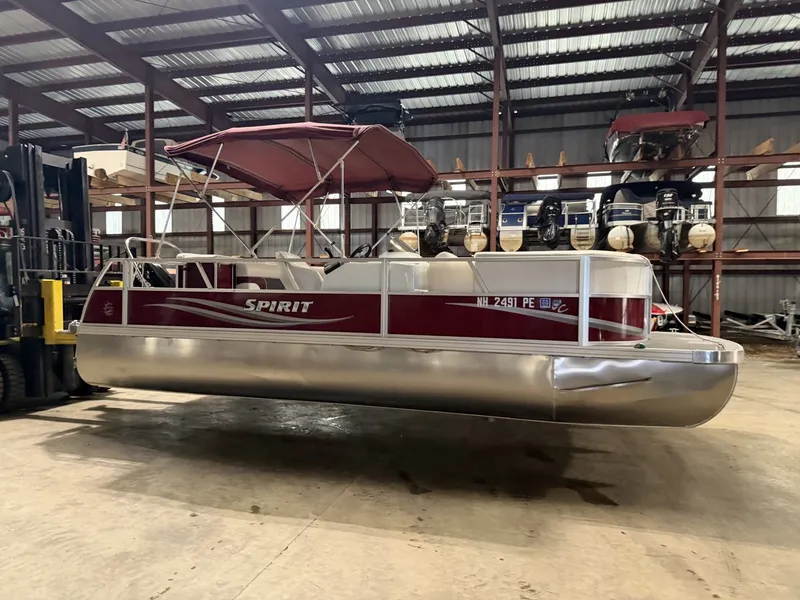 Slide: The Image of A 2011 JC Spirit pontoon boat in a warehouse, featuring a red canopy and silver hull. - 16