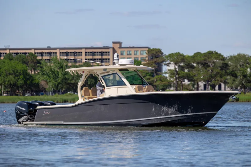 The Image of 2019 Scout 355 LXF boat cruising on a calm river with trees and buildings in the background. - 0