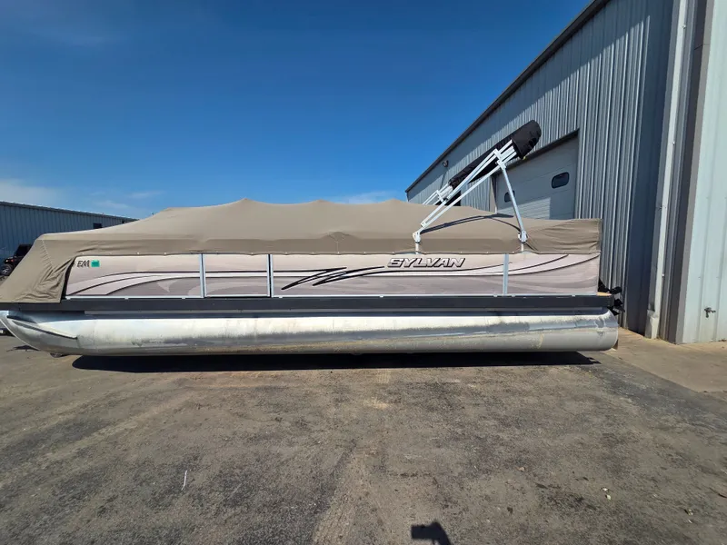 Slide: The Image of A 1998 Sylvan 826 Nexus pontoon boat with a cover, parked beside a building. - 2