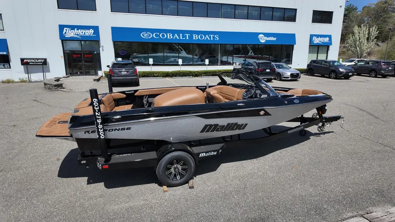 Slide: The Image of A 2025 Malibu TXi boat on a trailer in front of a boat dealership building. - 2