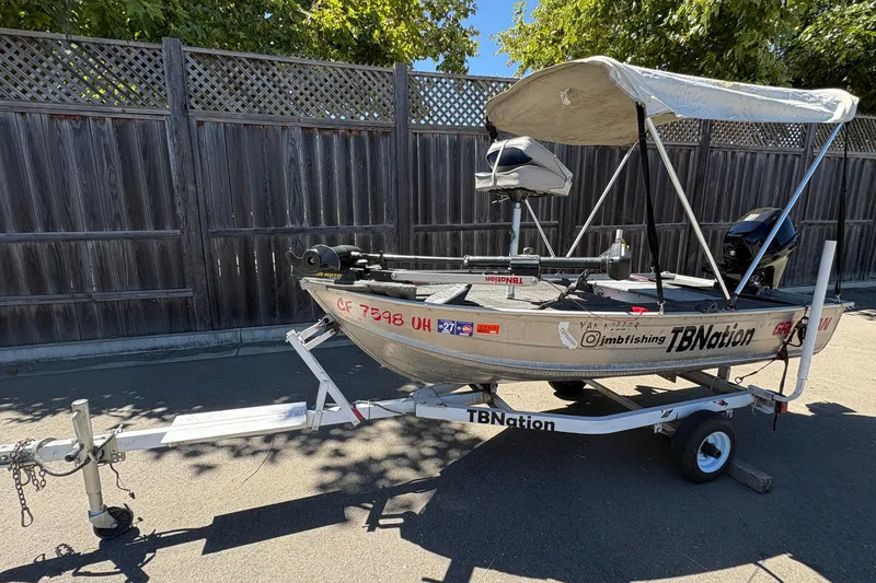 Slide: The Image of 1994 Grumman 1065 boat on trailer with canopy, parked by wooden fence. - 2