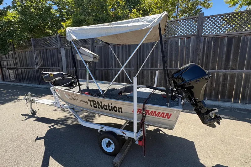 Slide: The Image of 1994 Grumman 1065 boat with canopy and motor on trailer, parked by wooden fence. - 3