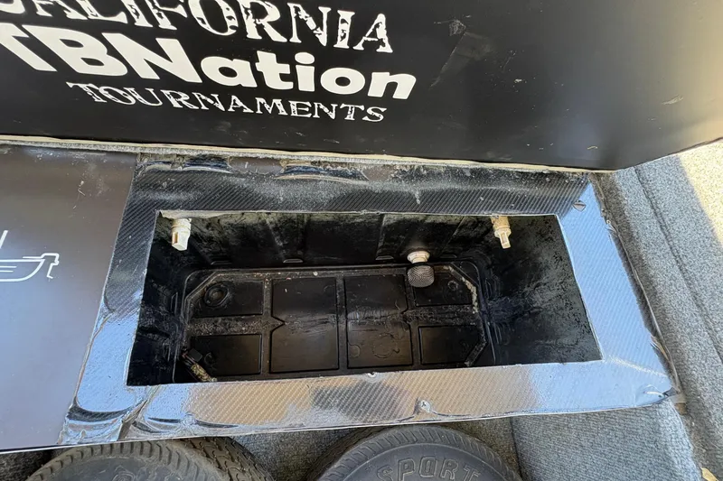 Slide: The Image of Open compartment in a 1994 Grumman 1065 boat, featuring storage space and visible tire. - 14