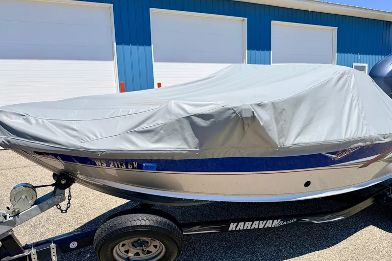 Slide: The Image of 2005 Alumacraft Tournament Sport 175 boat covered on trailer, parked near blue building. - 89