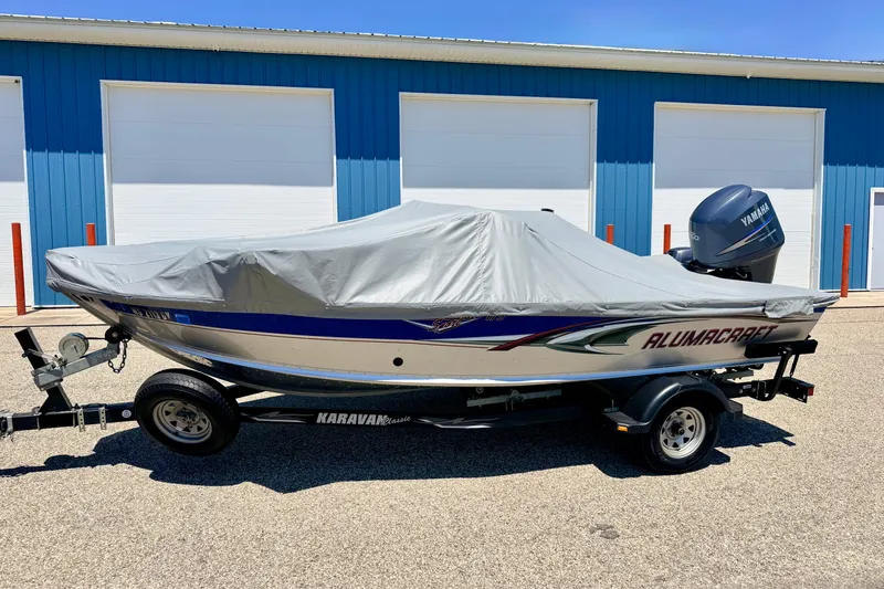 Slide: The Image of 2005 Alumacraft Tournament Sport 175 boat with cover on trailer, parked by blue building. - 87