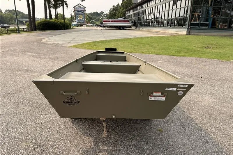 Slide: The Image of 2026 Tracker Grizzly 15 Jon boat on pavement near a showroom, showcasing its sturdy design. - 3