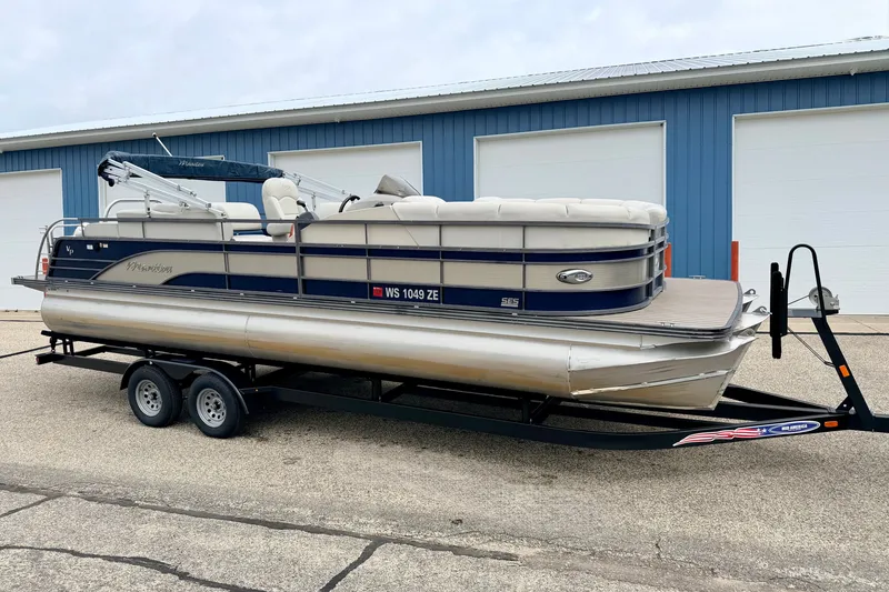 Slide: The Image of 2015 Manitou 25 SES BENCH pontoon boat on trailer, parked near blue building. - 2
