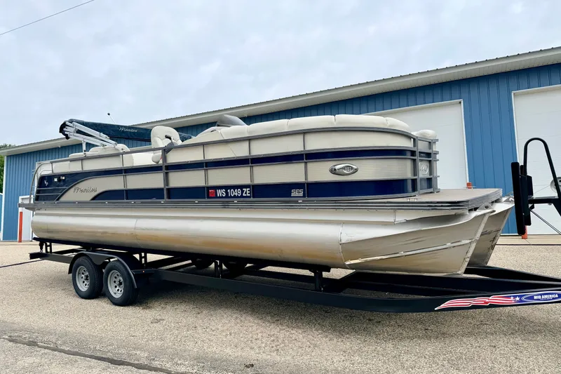 The Image of 2015 Manitou 25 SES BENCH pontoon boat on trailer, parked near blue building. - 0