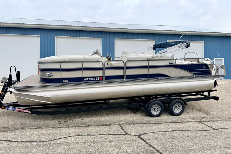 Slide: The Image of 2015 Manitou 25 SES BENCH pontoon boat on trailer, parked outside blue building. - 1