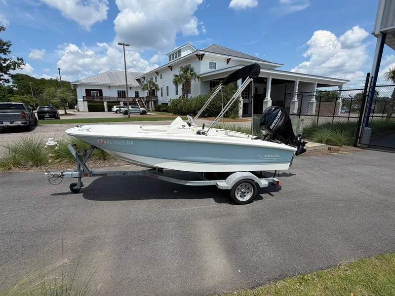 Slide: The Image of A 2018 Boston Whaler 130 Super Sport on a trailer in a parking lot. Buildings and trees are in the background. - 0
