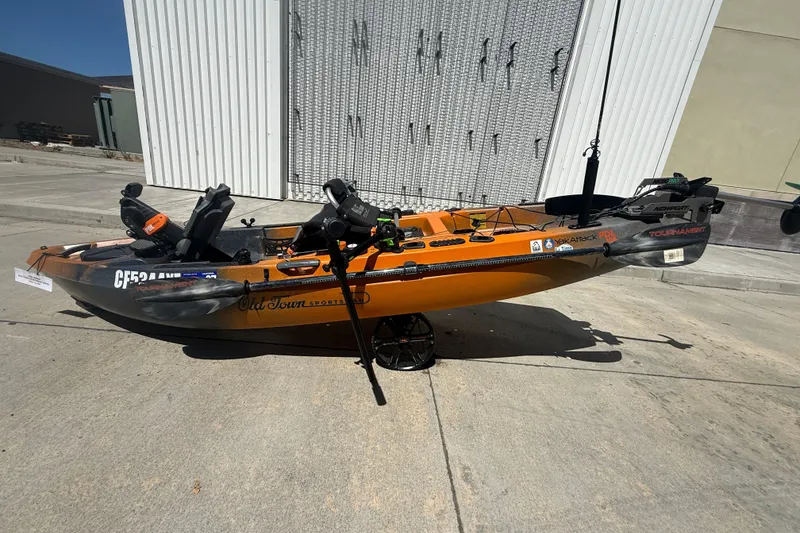Slide: The Image of Orange 2023 OldTown Sportsman PDL 106 kayak with fishing gear, parked outdoors. - 11
