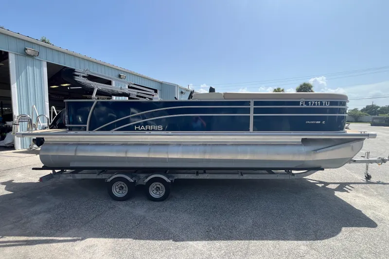 Slide: The Image of 2024 Harris Cruiser 230 pontoon boat on trailer, parked outside a blue building. - 4