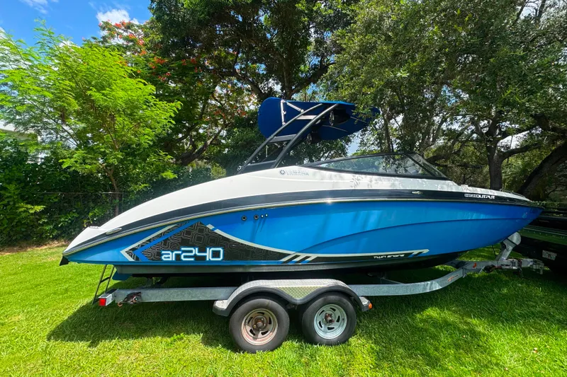 The Image of 2015 Yamaha Boats AR240 on trailer, blue and white, parked on grass under trees. - 0