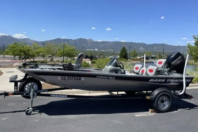 Slide: The Image of 2015 Tracker Bass boat on trailer, parked outdoors with scenic mountain backdrop. - 4