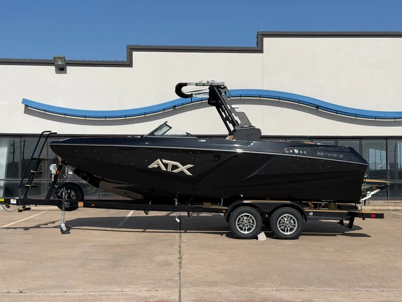 The Image of A black ATX Surf Boat, model 22 Type-S, is on a trailer in front of a modern building. - 1