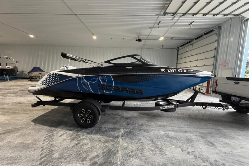 The Image of 2022 Scarab 195 ID boat in a garage, blue and black design, on a trailer. - 0
