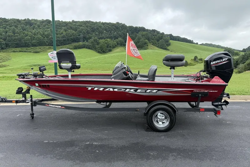 The Image of 2024 Tracker Pro Team 175 TF boat on trailer, red color, parked on asphalt with green hills background. - 1