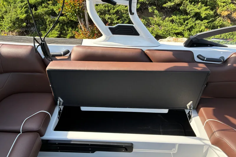 Slide: The Image of 2025 Moomba Max boat interior with open storage compartment, brown seating, and lush greenery background. - 24