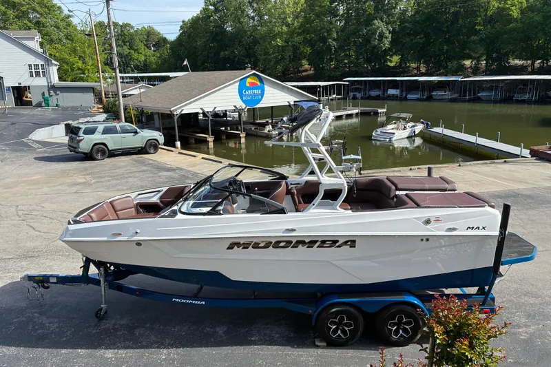 Slide: The Image of 2025 Moomba Max boat on trailer near marina, surrounded by trees and parked vehicles. - 7