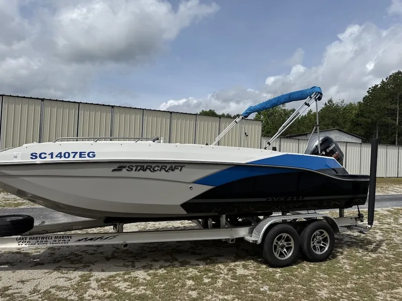 The Image of A 2023 Starcraft SVX 211 OB boat on a trailer, featuring blue and black design, near a building. - 1