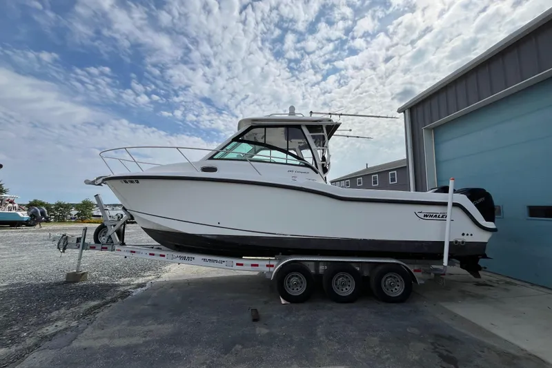 Slide: The Image of 2012 Boston Whaler 285 Conquest boat on trailer, parked near a blue garage under a cloudy sky. - 2