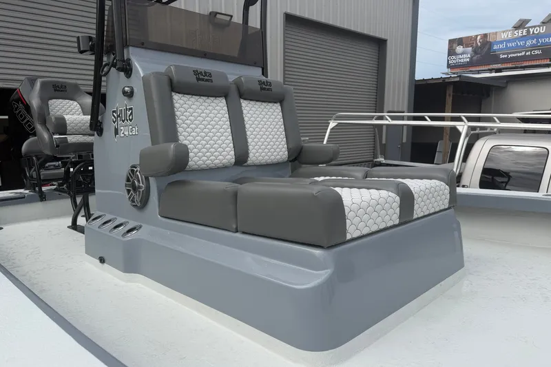Slide: The Image of 2027 Skuta 24 Cat boat with gray and white cushioned seating, docked near a warehouse. - 5