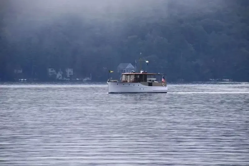 Slide: The Image of A classic 1926 boat cruises on a calm lake, surrounded by wooded hills. - 5