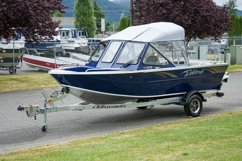 Slide: The Image of 2027 Weldcraft 180 Rebel boat on trailer, parked outdoors with trees in background. - 22