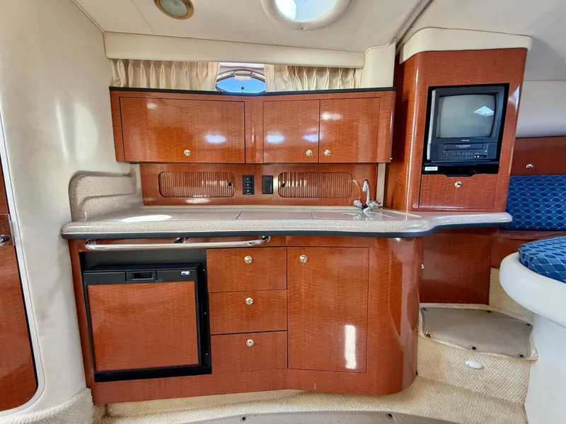 Slide: The Image of The interior of a 2003 Sea Ray Sundancer 320 features an elegant galley with wood cabinets, a small fridge, and a built-in TV. - 13