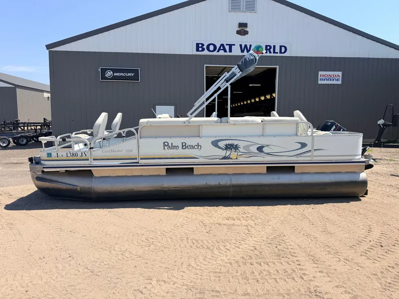 Slide: The Image of A 2004 Palm Beach CastMaster 200 pontoon boat with a 50HP Mercury 4Stroke engine, parked on sandy ground in front of a building labeled "Boat World." - 28