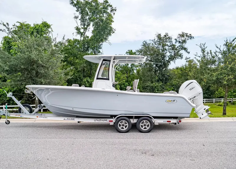 Slide: The Image of A 2027 Sea Hunt Ultra 245 SE boat on a trailer on a road, surrounded by trees. - 2