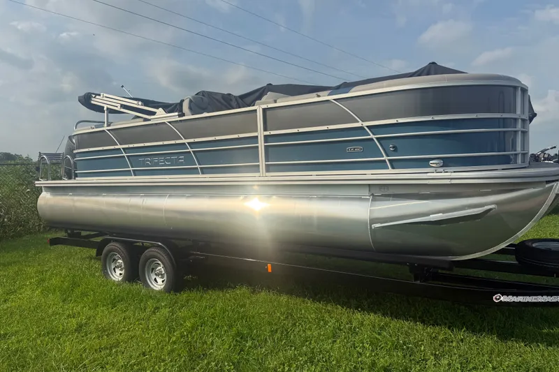 Slide: The Image of 2025 Trifecta 24 RF LE pontoon boat on trailer, parked on grass under cloudy sky. - 2