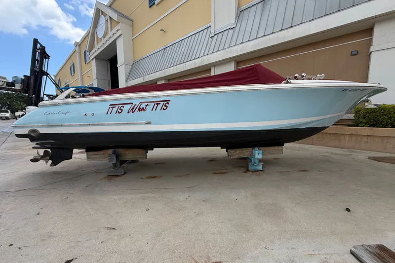 Slide: The Image of 2005 Chris-Craft Corsair 25 boat on blocks, light blue hull, red cover, outdoor setting. - 1