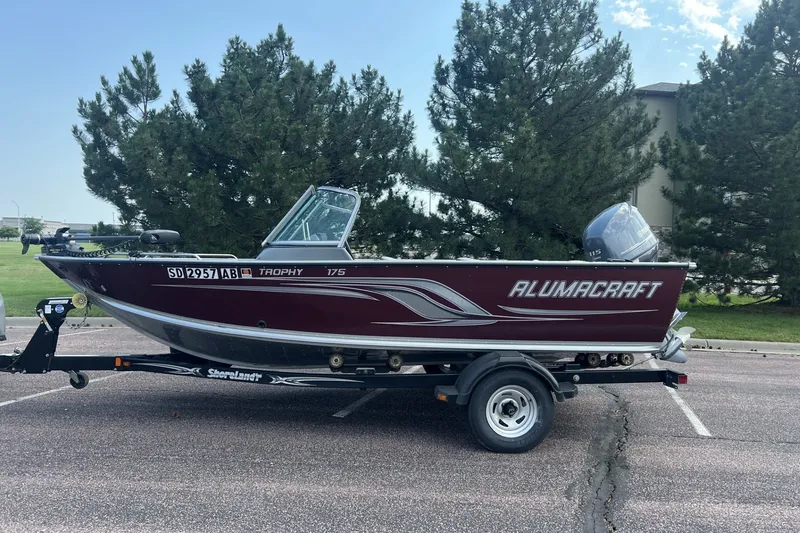 The Image of 2012 Alumacraft Trophy 175 boat on trailer, parked outdoors with trees in background. - 1